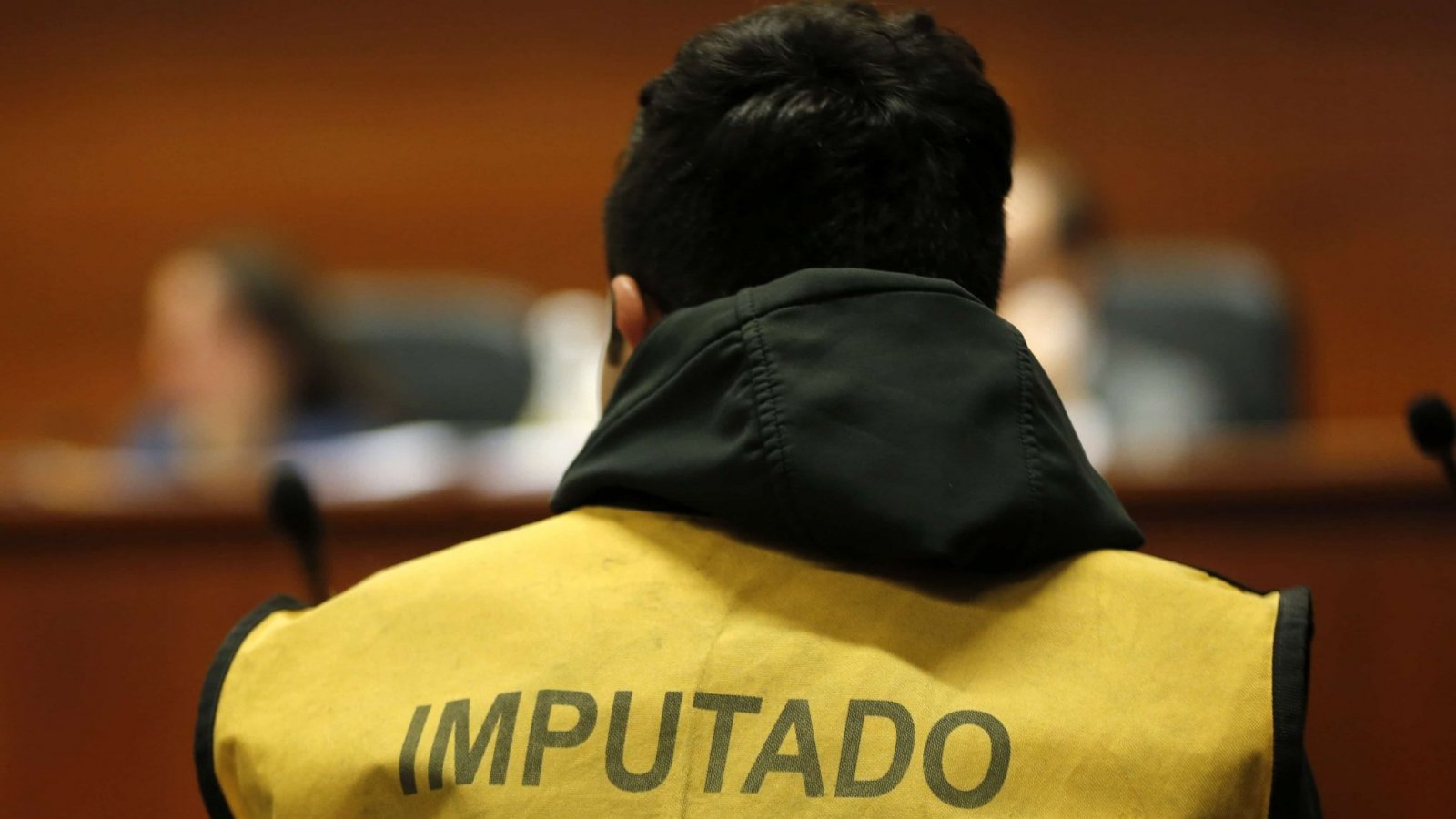 Imagen referencial de la espalda de un hombre en un tribunal, vistiendo una chaqueta amarilla que dice "imputado".