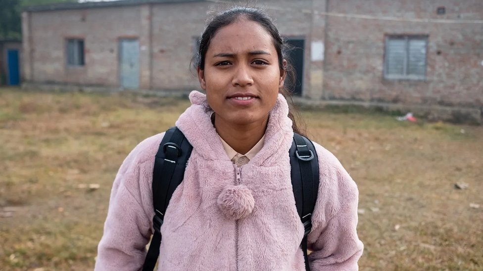 Una joven de 18 años, con sudadera de peluche rosado y mochila, posa delante de una pequeña escuela de ladrillos.