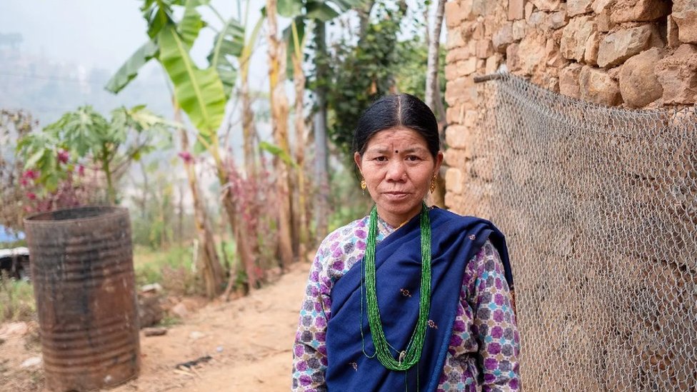 Una mujer mayor con vestido y oga azul, un largo collar verde, pendientes de oro y un bindi en la frente, posa delante de un pequeño jardín de plataneras.