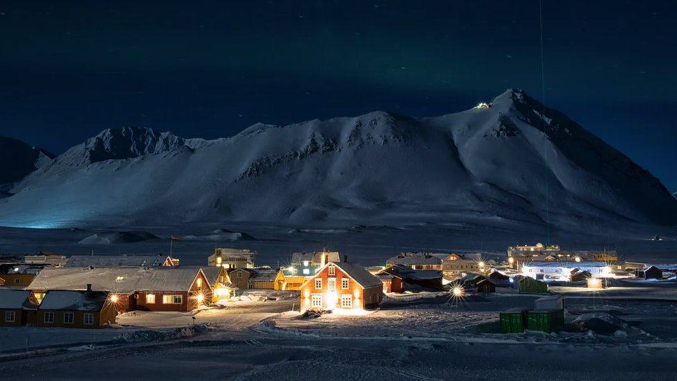 Diminuto asentamiento de la ciudad de Ny-Ålesund, - con las luces encendidas mientras la oscuridad cae sobre las montañas en el fondo