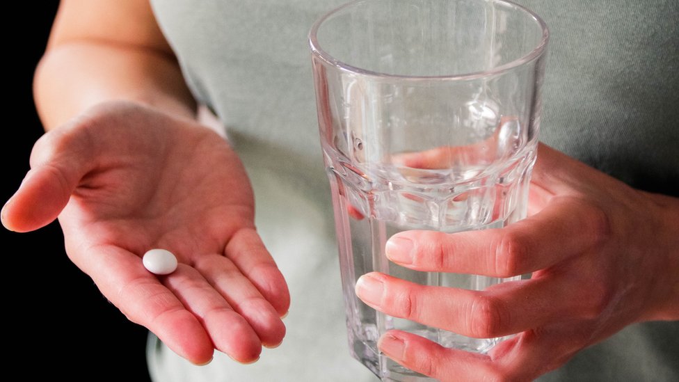 Una mujer sosteniendo una píldora en una mano y un vaso de agua en otra