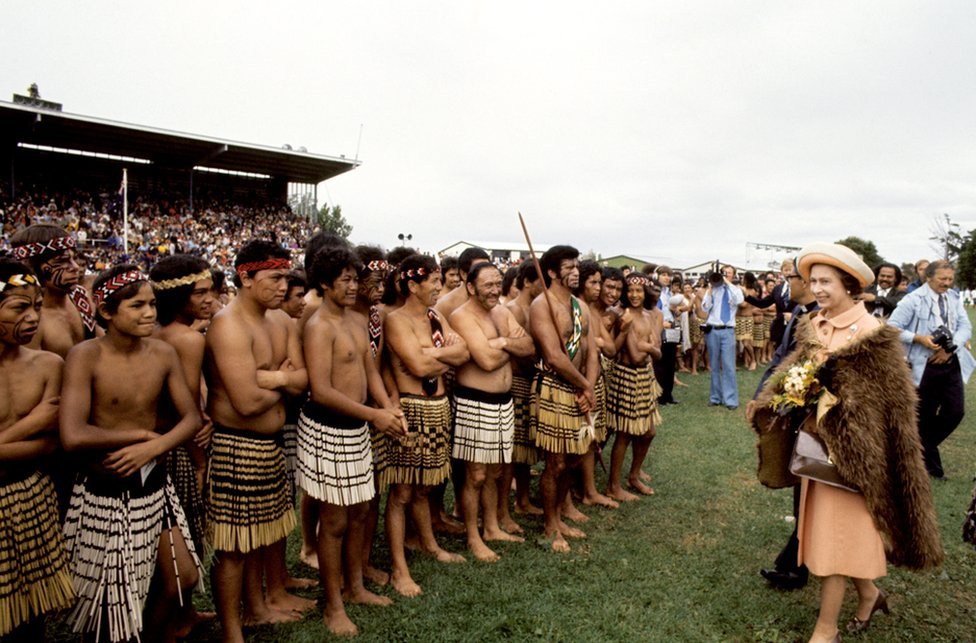 La reina Isabel II, con un manto de plumas marrones de kiwi, con guerreros maoríes en Rugby Park, Gisborne, cuando ella y el duque de Edimburgo recibieron una bienvenida maorí de Nueva Zelanda en la inauguración del Festival Polinesio Real de Nueva Zelanda.