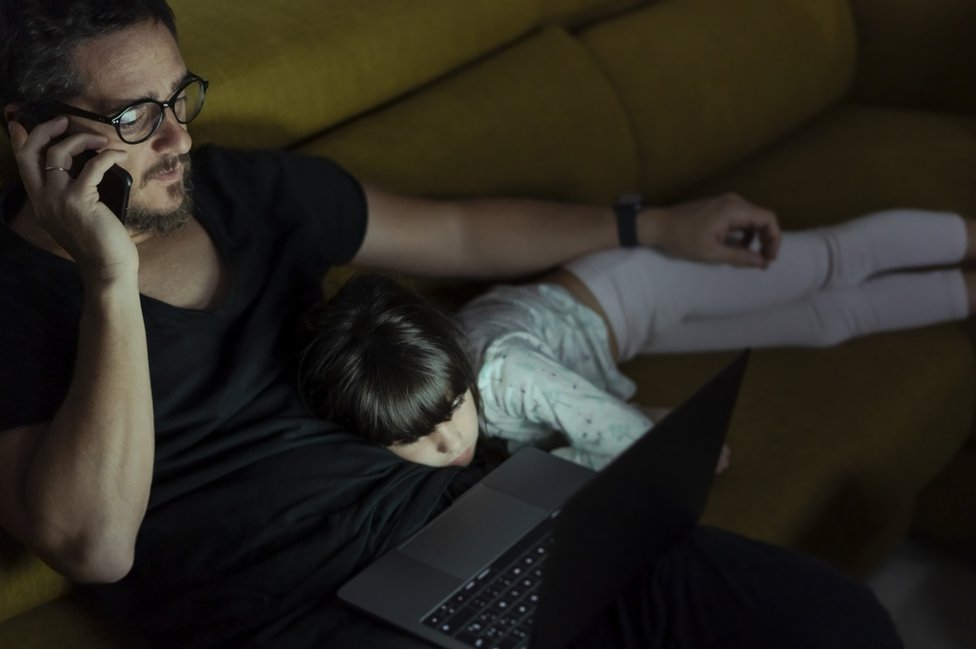 Un hombre teletrabajando hasta tarde con su niña acostada sobre sus piernas.