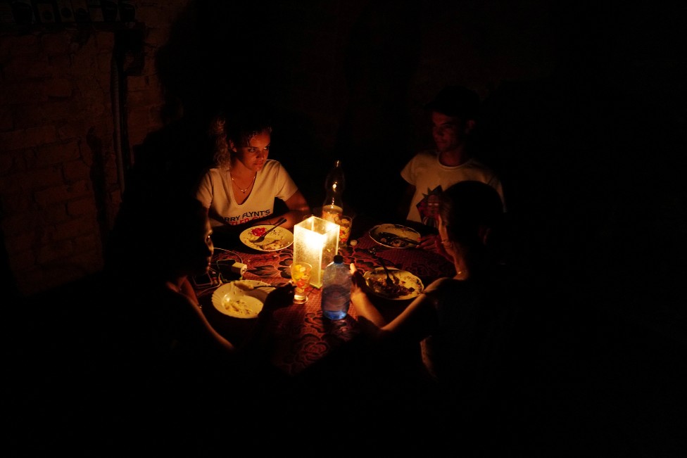 Tres personas de una familia sentdas a la mesa, todo está a oscuras y se alumbran con velas.