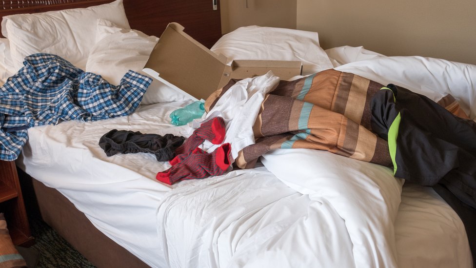 Una cama desarreglada con ropa, una botella de plástico y una caja de pizza encima.