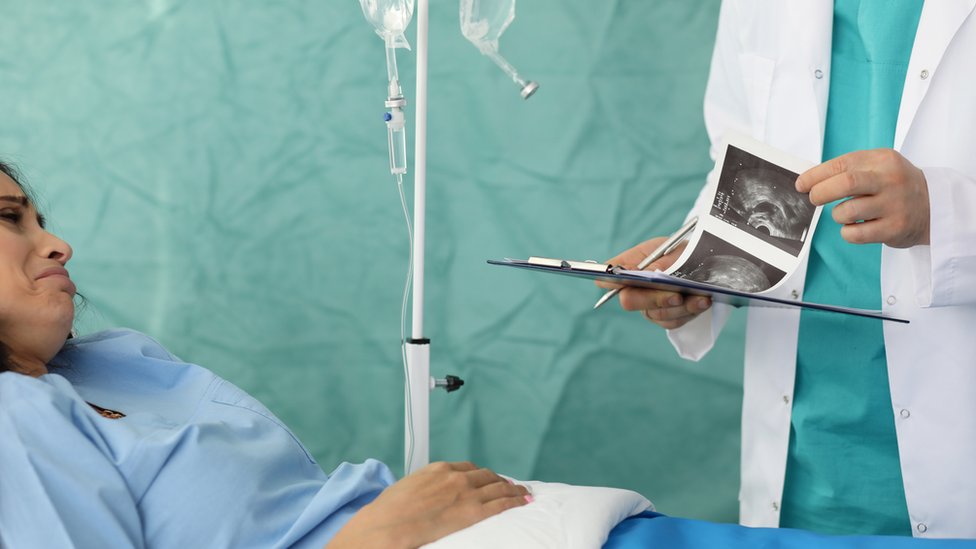 Una mujer en una cama de hospital reacciona con tristeza a la evaluación de un médico que mira una ecografía