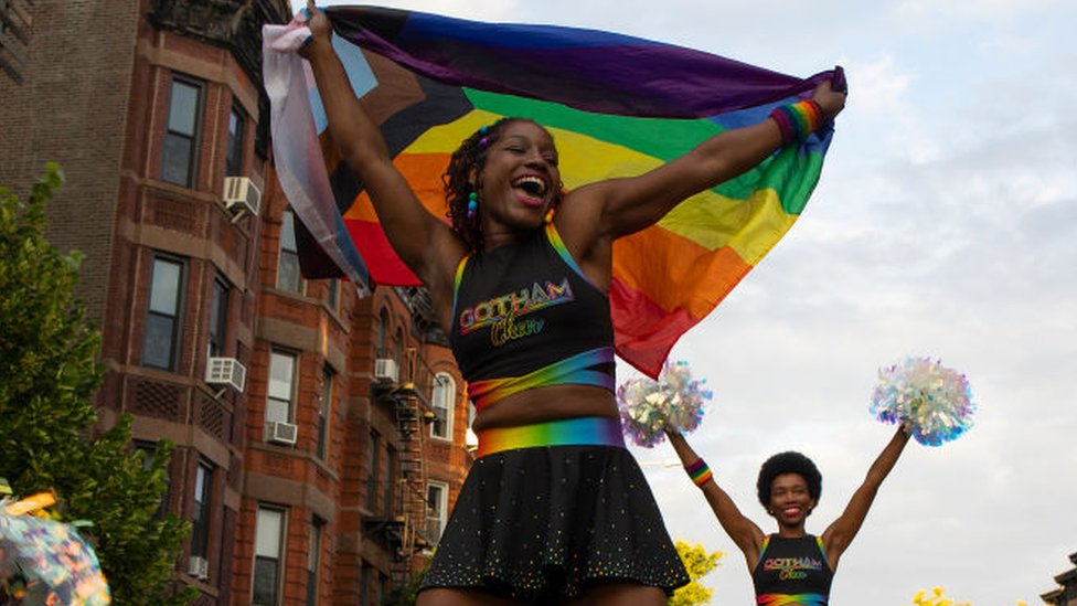 Mujer vestida de porrista con una bandera LGBT en sus manos.