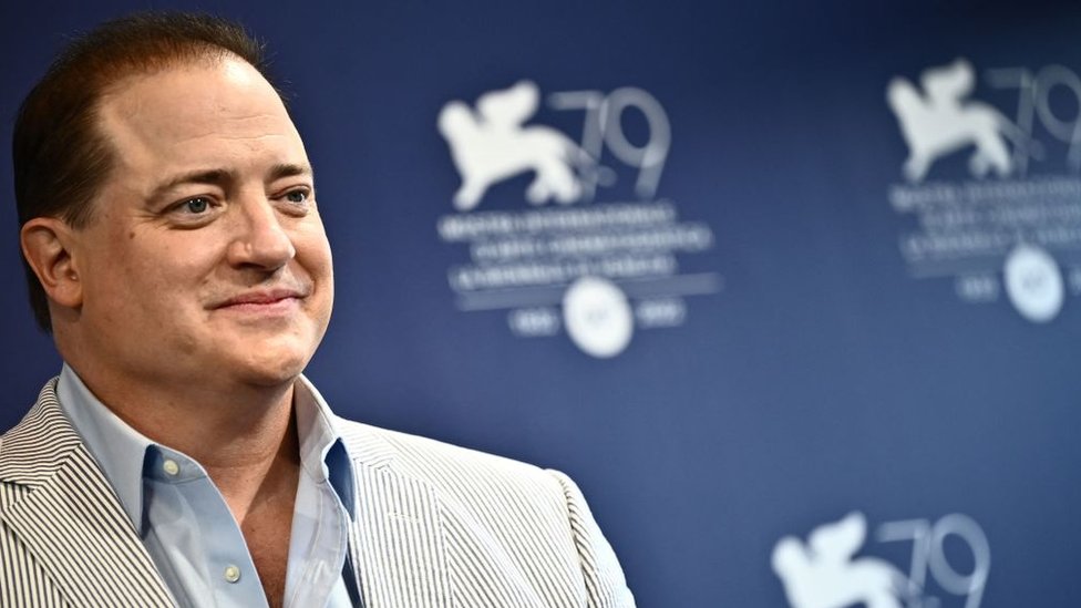 El actor Brendan Fraser vestido de traje claro con el fondo azul del cartel del Festival de Venecia.