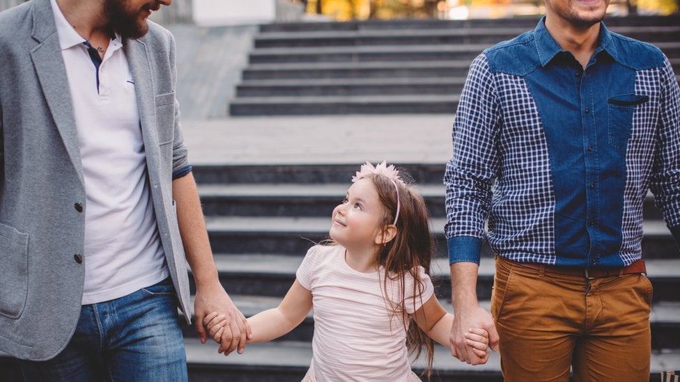 Una niña llevada de la mano por sus dos padres.