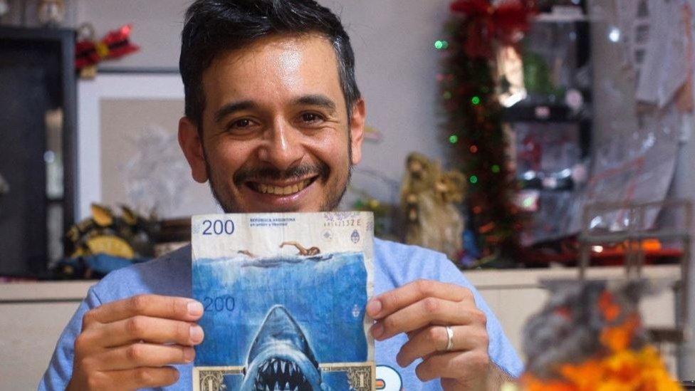 El artista Sergio Días sostiene una pintura hecha sobre billetes que incluye la imagen del tiburón de la película de Steven Spielberg