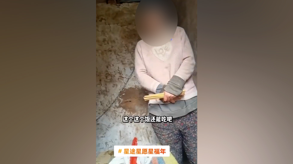 Una captura de pantalla del video muestra a la mujer con una cadena al cuello de pie en la cabaña