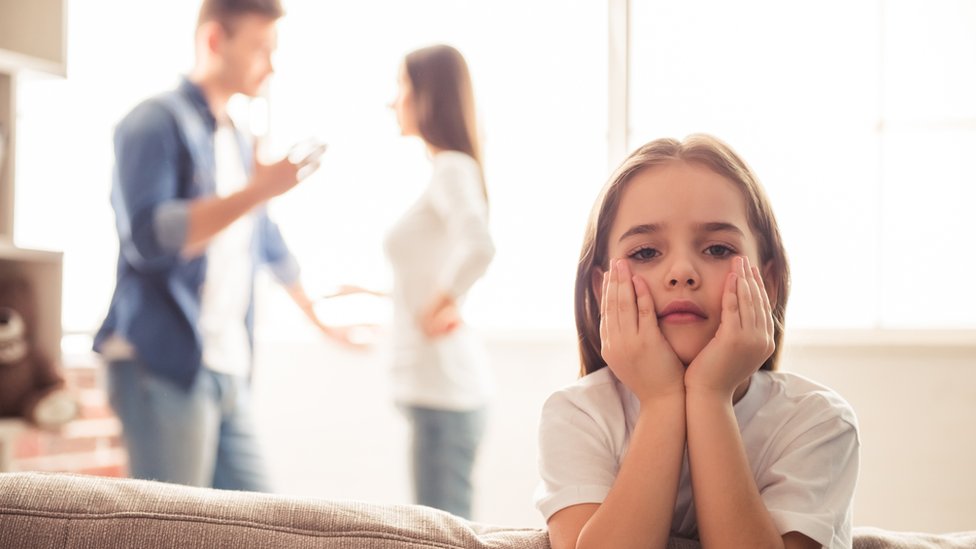 Una pareja discutiendo delante de una niña que mira a la cámara