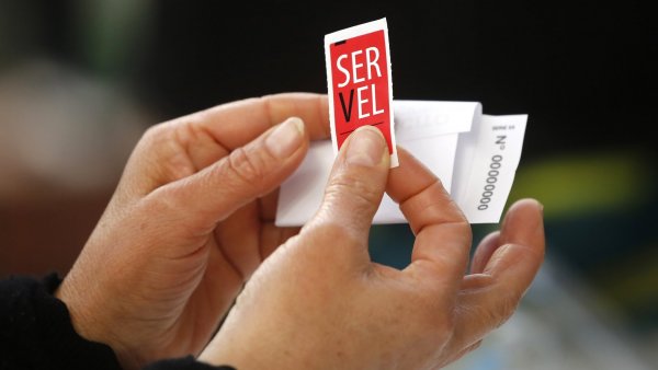 Manos sujetando un sticker del Servel y un voto doblado.-