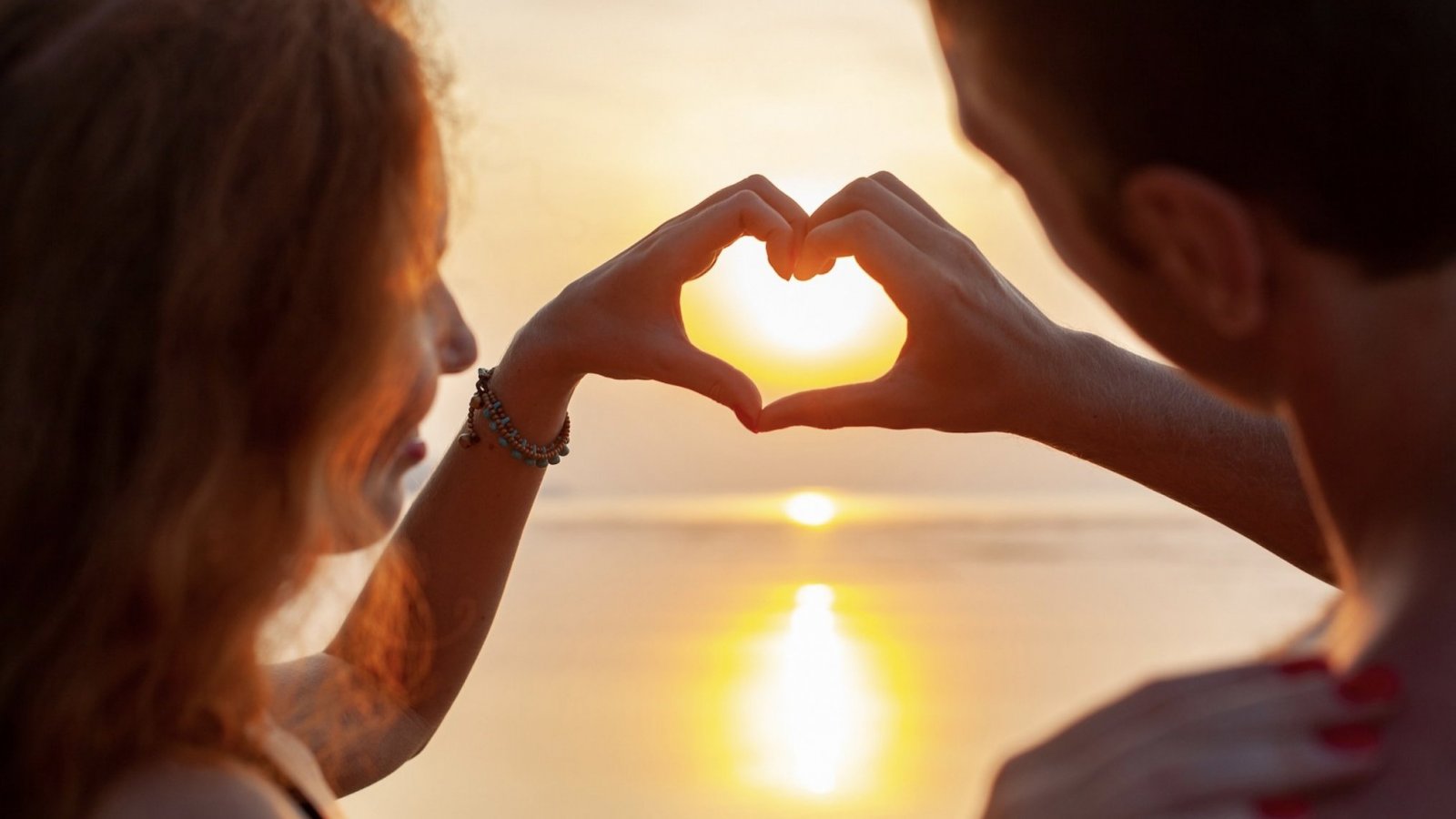 Una pareja frente a un atardecer haciendo un corazón con las manos.