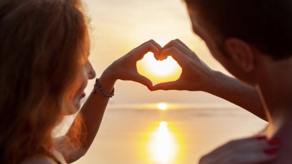 Una pareja frente a un atardecer haciendo un corazón con las manos.