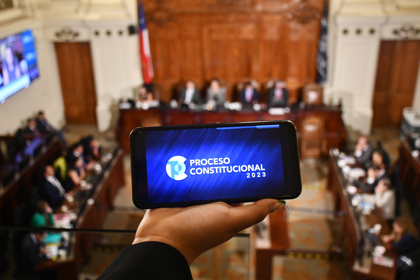 Celular con proceso constituyente en pantalla y de fondo la Comision Experta.