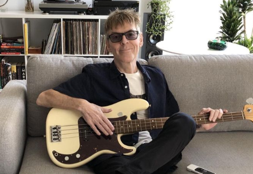 Andy Rourke sentado en un sofá con un bajo en sus manos.