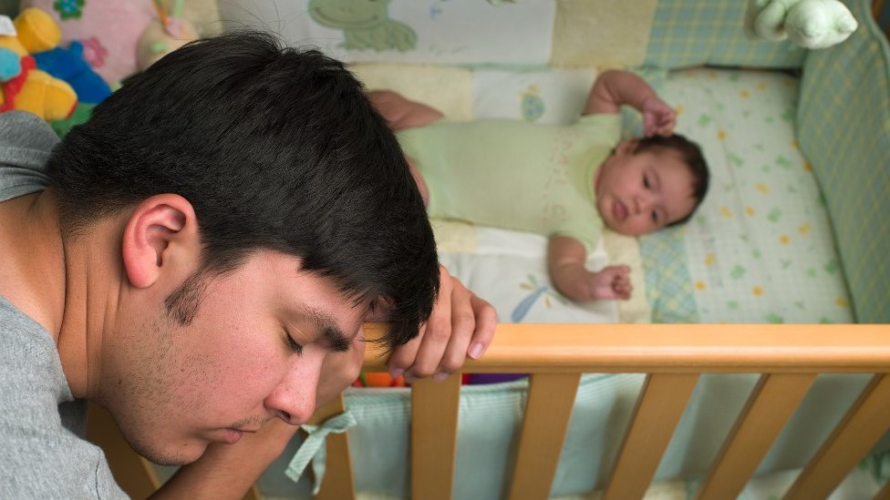Un hombre apoya la cabeza en la cuna junto a su bebé recién nacido.