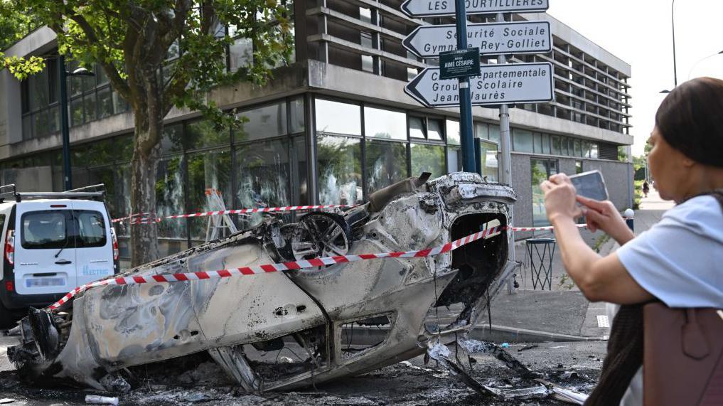 Esta fotografía tomada el 30 de junio de 2023 muestra a un peatón fotografiando un automóvil incendiado volcado en Aubervilliers, al norte de París