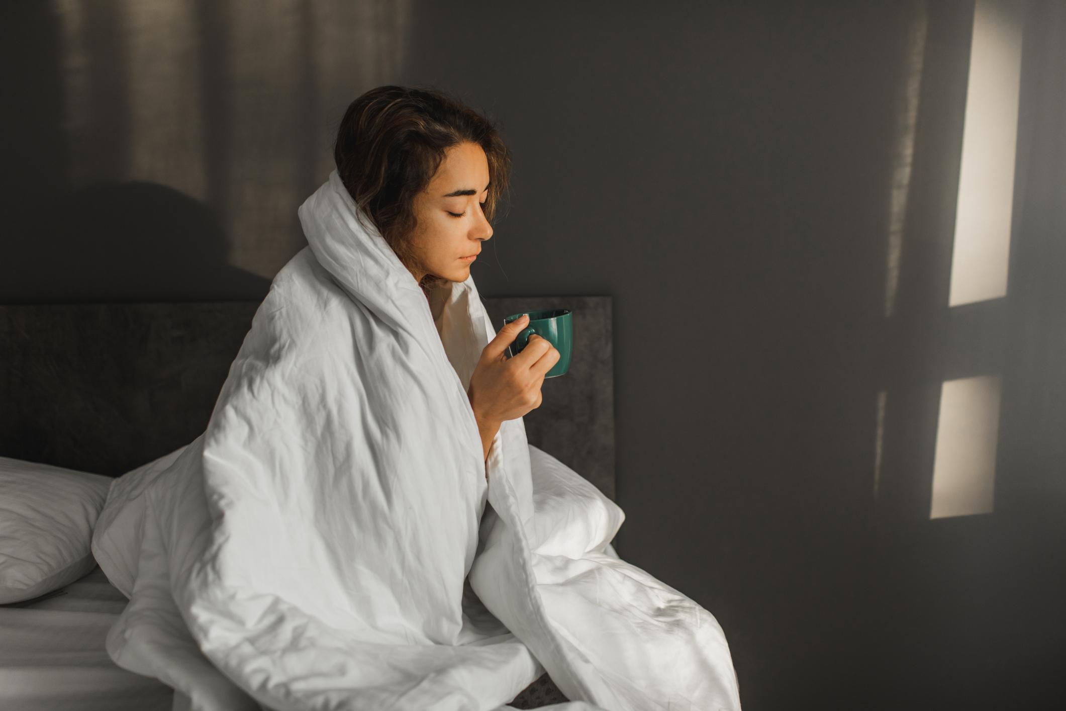 Una mujer enferma tiene fiebre y bebe té caliente en la cama y cubierta con una manta. 