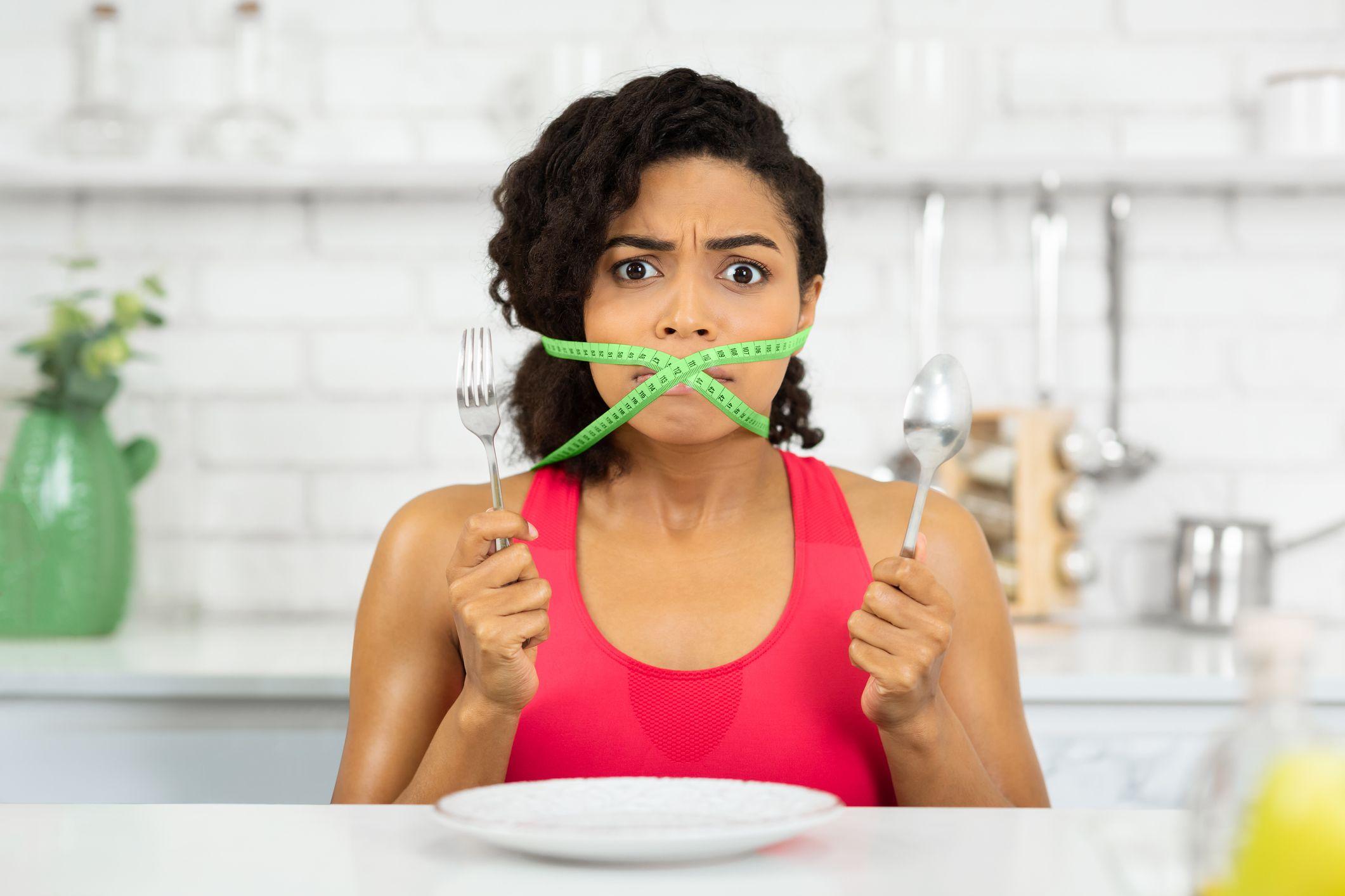 Mujer con una cinta métrica alrededor de la cabeza a la altura de la boca, con un tenedor y una cuchara en cada mano.