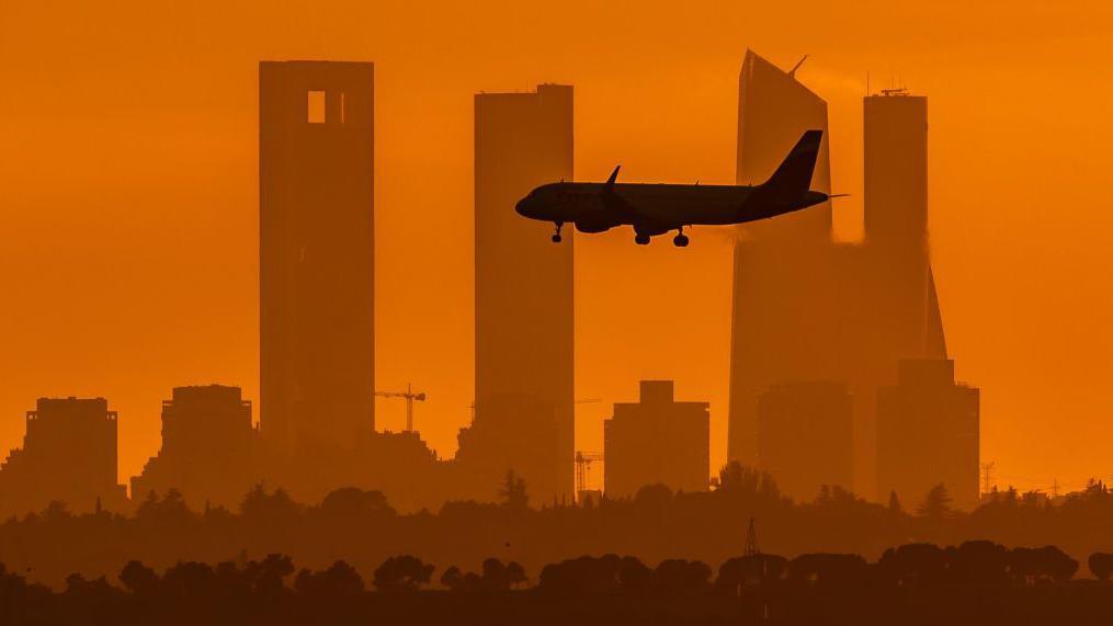 Un avión con las torres de Madrid de fondo y el cielo teñido de naranja