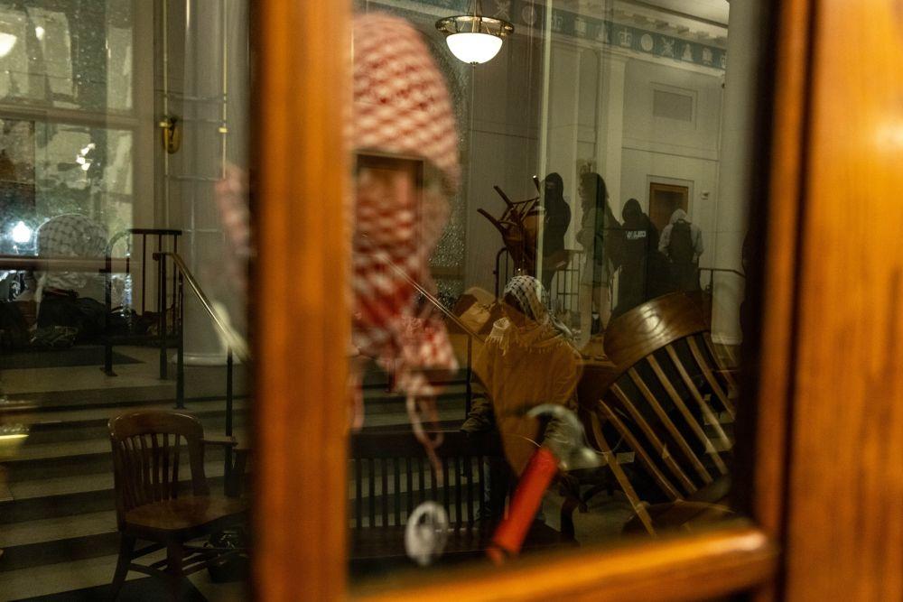 Manifestantes cargando con sillas mientras otro mira por la ventana con un martillo en la mano.