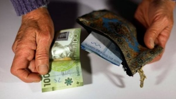 Dos ancianos alrededor de un frasco lleno de billetes, con las letras 