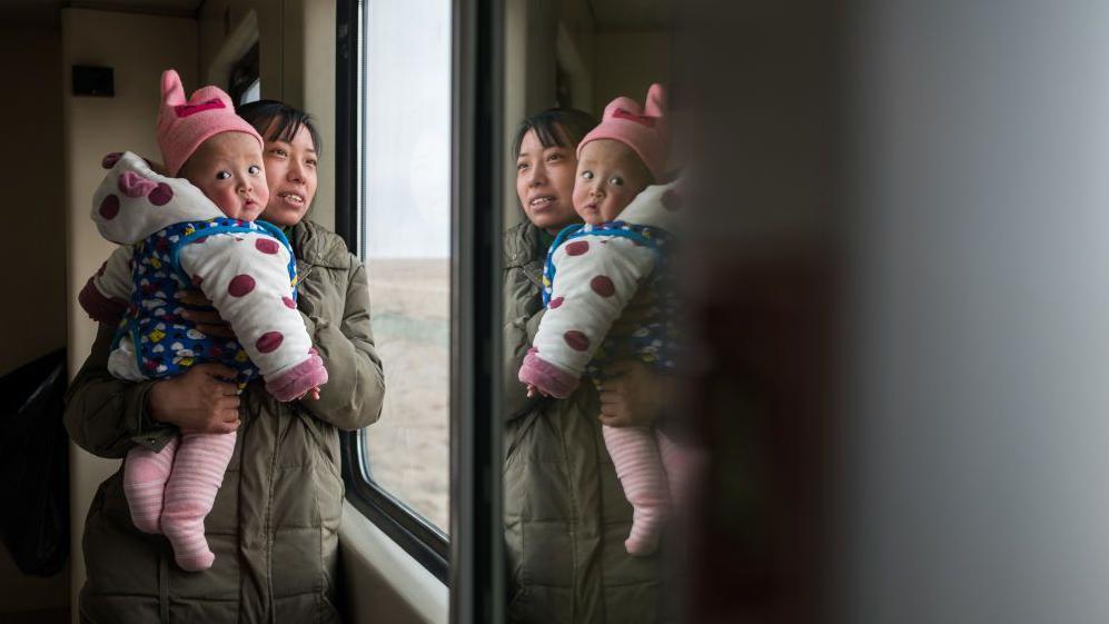 Una china y carga a su bebé en brazos junto a la ventana de un tren.