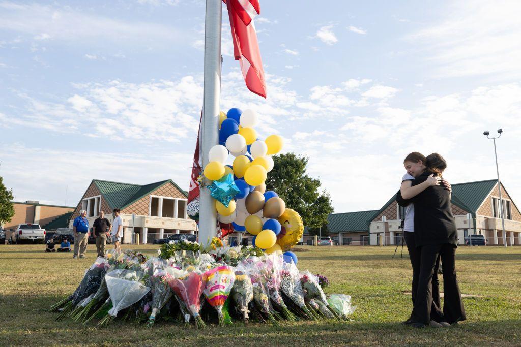 Dos adolescentes se abrazan al lado de un lugar lleno de flores que recuerdan a las víctimas del tiroteo.