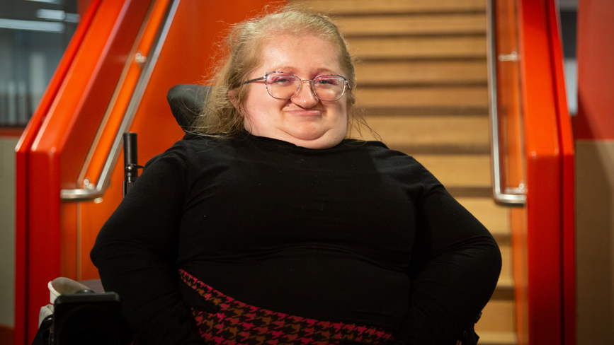 Una mujer en silla de ruedas sonríe a la cámara. Lleva un top negro con una falda negra y roja de pata de gallo. Detrás de ella hay unas escaleras con las barandillas pintadas de naranja.