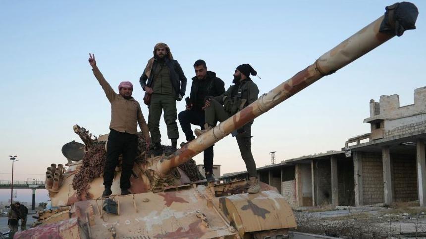 Cuatro hombres, en pie sobre un tanque abandonado en la carretera que conduce a Alepo. Uno de ellos hace un gesto de victoria.