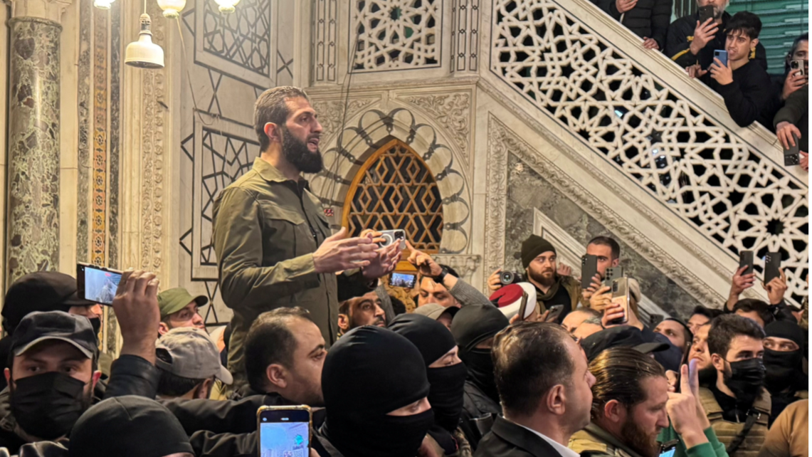 Abu Mohamed al Jolani, vistiendo una chaqueta verde oliva, de pie frente un grupo de hombres dentro de una mezquita.