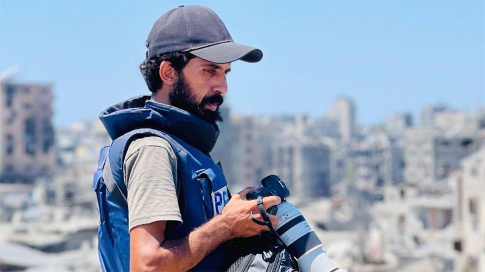 Un hombre con barba y gorra de béisbol sostiene una cámara . Está de perfil y lleva un chaleco antibalas con la palabra "Prensa". Al fondo se ve una imagen borrosa de lo que parece ser una ciudad fuertemente bombardeada.
