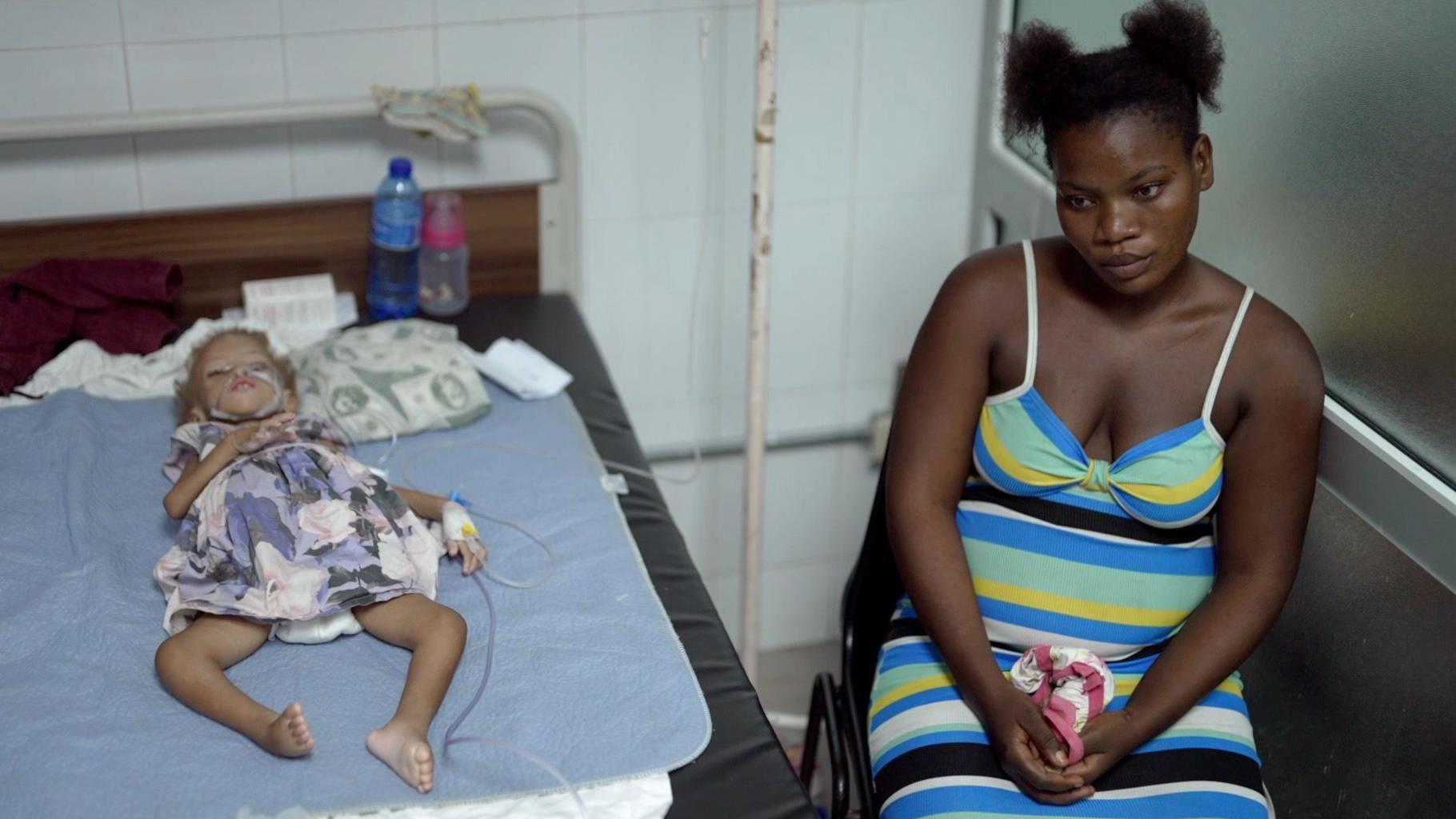 Una niña pequeña y demacrada yace en una cama conectada a un gotero mientras su madre está sentada a su lado. La madre sostiene un paño en sus manos.