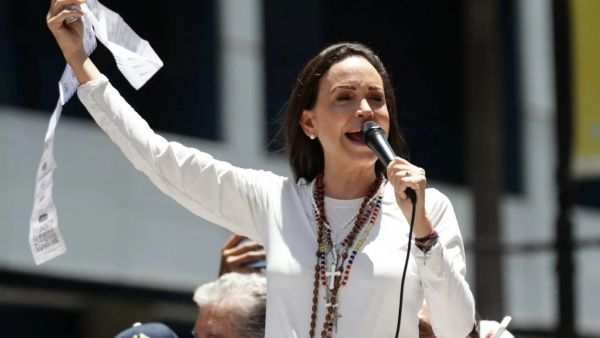 María Corina Machado, vestida de blanco, hablando en público con un micrófono en la mano izquierda y un papel en la mano derecha