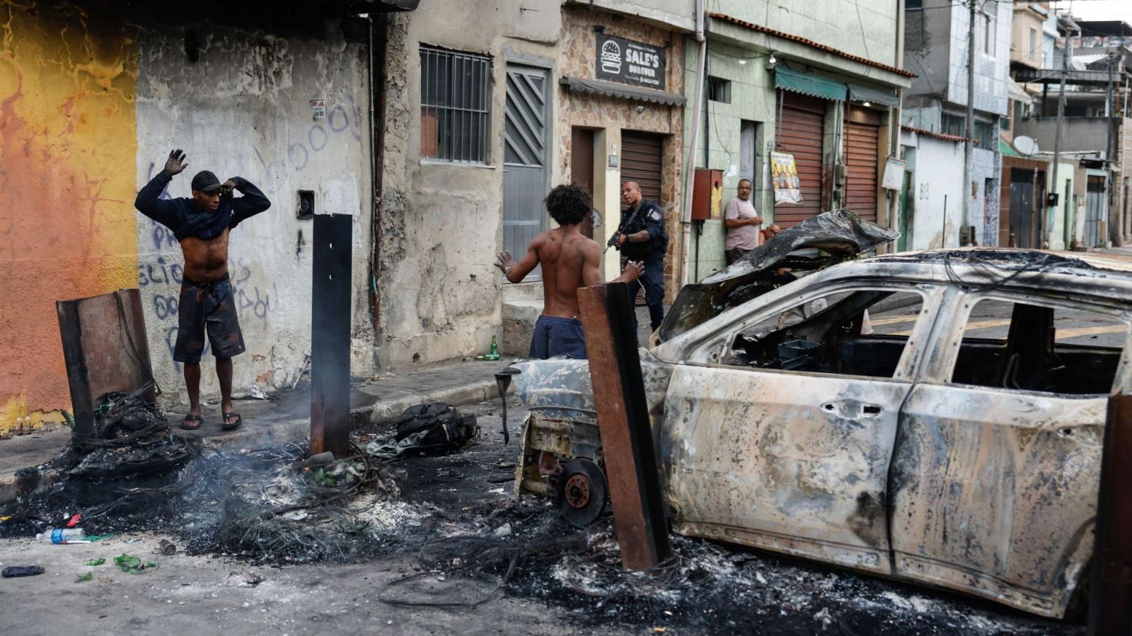 Policía con arma en la calle, auto quemado y hombres con los brazos arriba en medio de operativo.